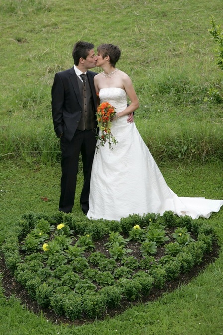 Hochzeitspaar küssst sich auf wiese in hanglage, vor ihnen ein aus pflanzen zusammengesetztes herz