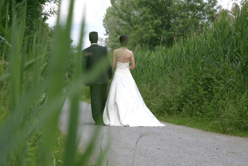 brautpaar von hinten aufgenommen, sie laufen auf einer kleinen strasse zwischen schilfgürtel vom fotografen weg