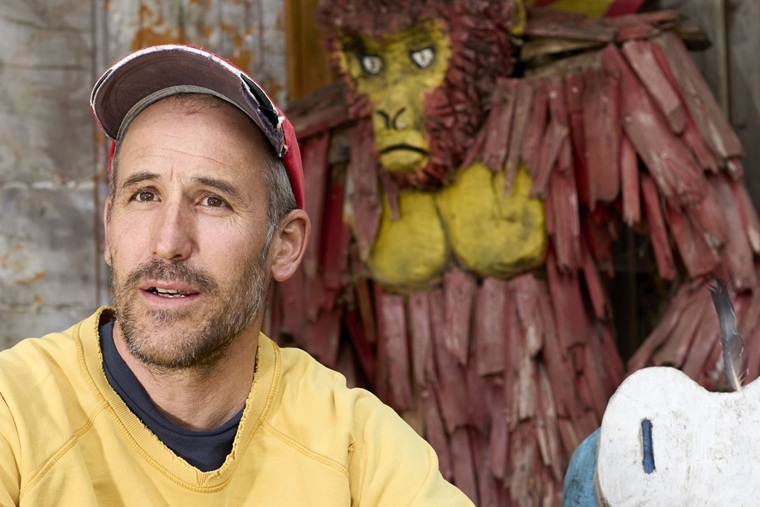 Künstler Leto mit gelbem Sweater und Cap vor Holzskulptur