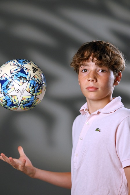 junge mit rosa polo-shirt wirft fussball in die höhe und schaut in die kamera